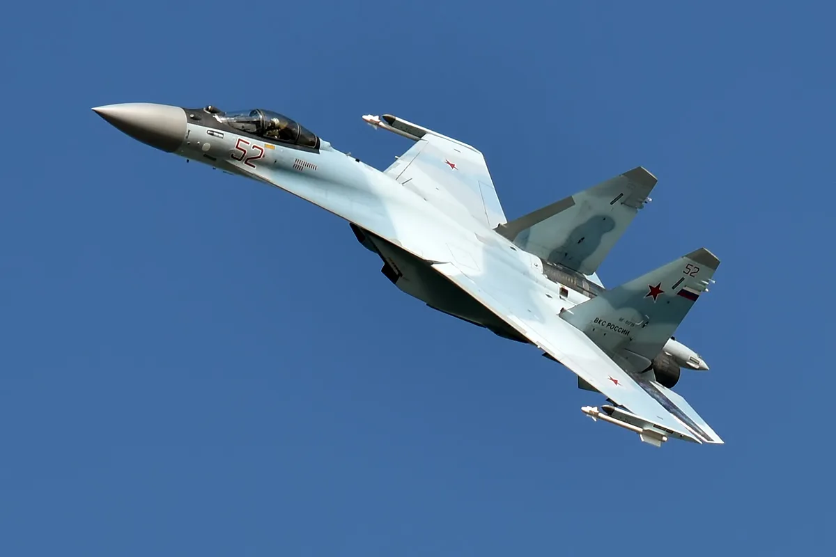 A Su-35S fighter jet banking sharply against a clear blue sky, full side profile.