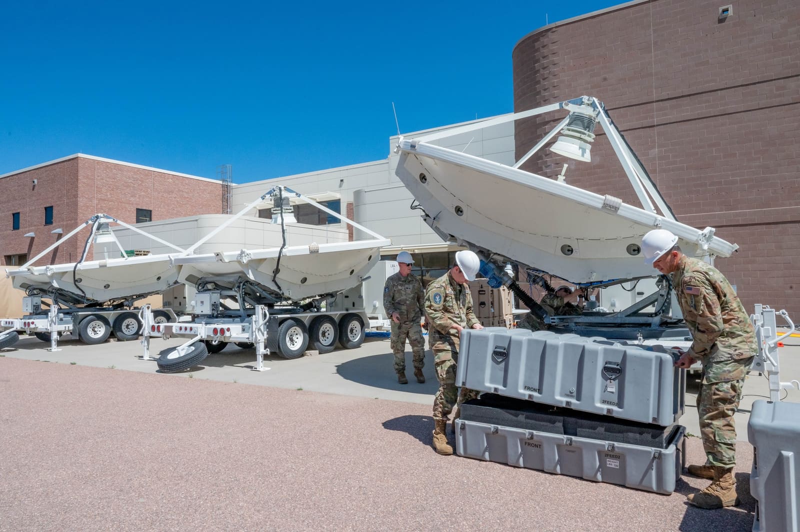 Multiple trailer-mounted Counter Communications System dishes lined up outside a building at Peterson Space Force Base, with Guardians in white hard hats working around shipping cases in the foreground