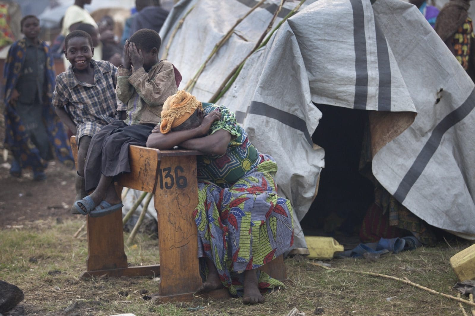 A woman and children sit beside a wooden bench and makeshift tent shelter in a displaced-persons camp