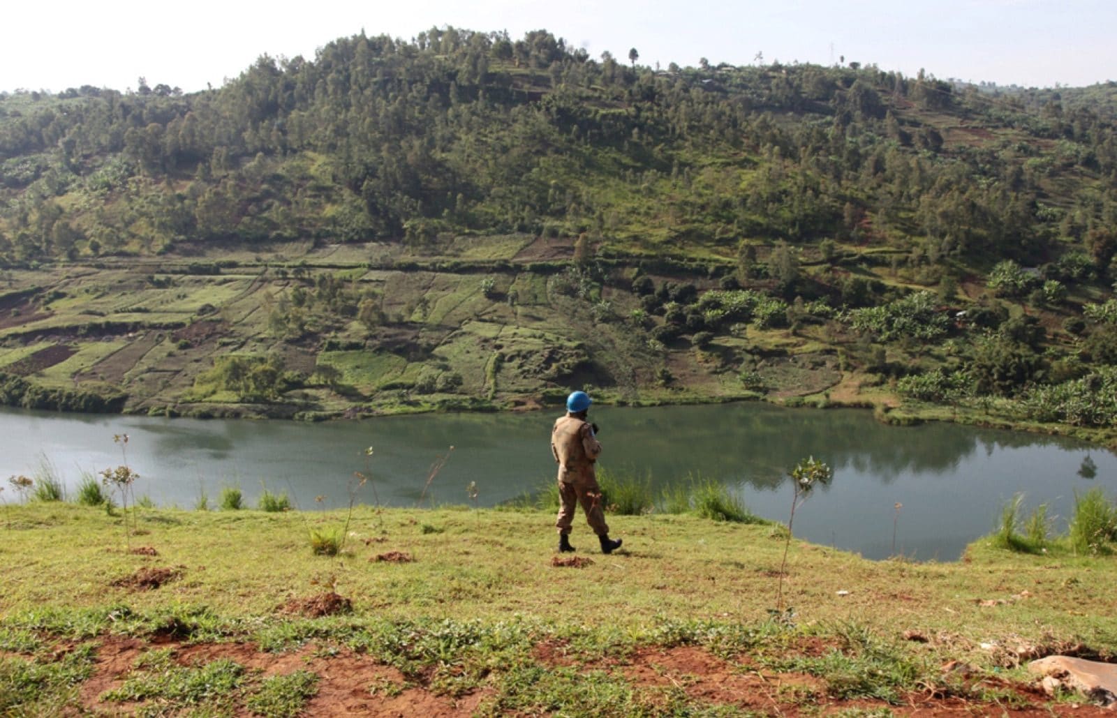 A blue-helmeted MONUSCO peacekeeper walks along the grassy shore of Lake Kivu in Goma, North Kivu, with hills and farmland visible across the water
