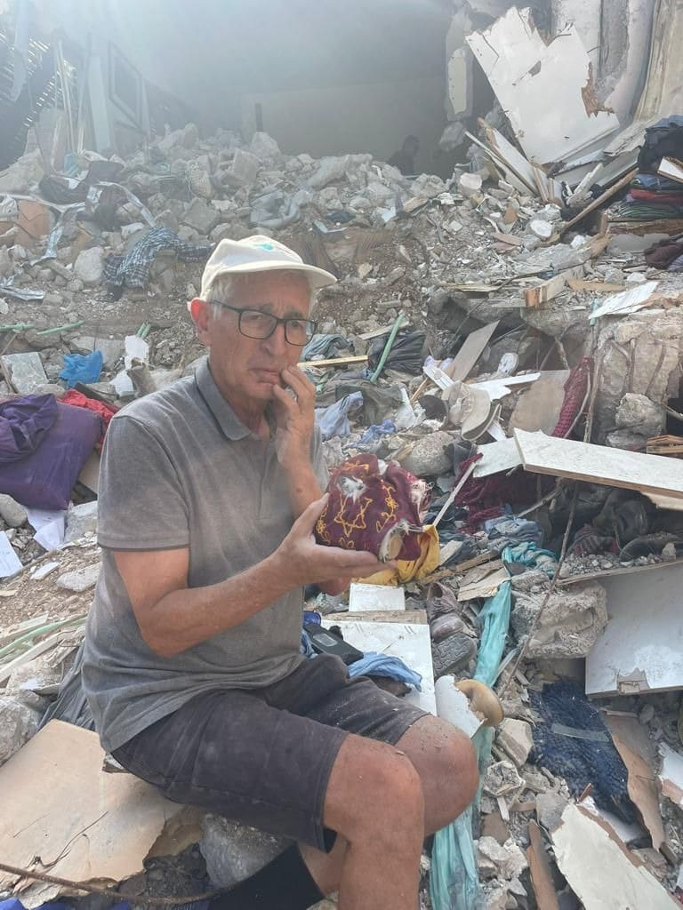A man, Shai Hermesh, stands amid the burned ruins of his home in Kibbutz Kfar Aza, holding tefillin he recovered from the wreckage
