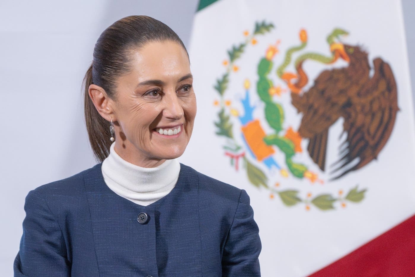 Claudia Sheinbaum smiling at her morning press conference, in front of Mexico's national coat of arms, announcing the tariff pause